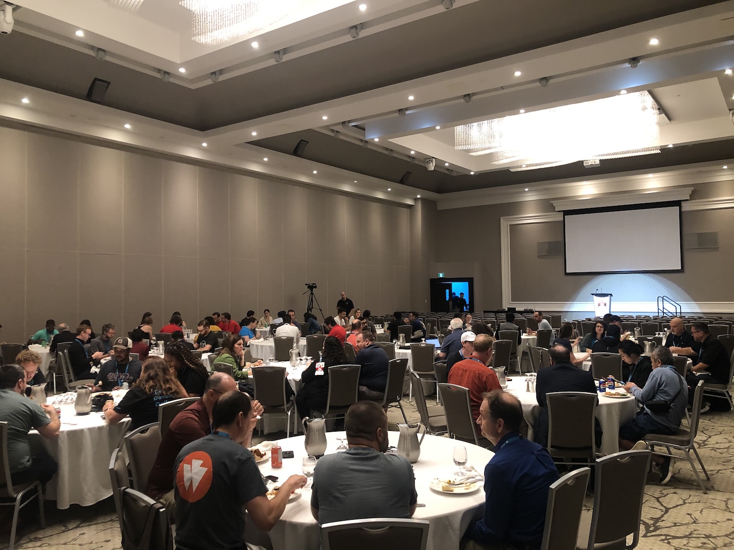 A crowd eating in a large conference room. Round tables, with white tablecloths. A stage with a podium lit in white light is set up at the far side of the room.